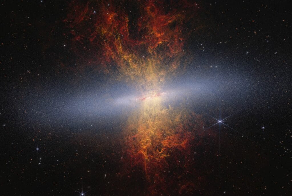 Kolorowe zdjęcie kompozytowe M82 („Galaktyki Cygaro”) z Webba i Hubble’a: cienki, krawędziowo widziany dysk, w którym Webb ujawnia drobnoziarnistą strukturę gwiazdową, oraz jasne, czerwone pióropusze gazu wystrzeliwujące ponad i poniżej płaszczyzny galaktyki
