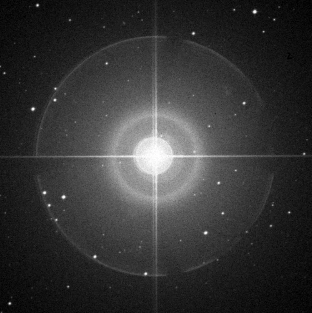 Czarno-białe archiwalne zdjęcie nieba z przeglądu Digitized Sky Survey, przedstawiające pole gwiazd o różnej jasności na ciemnym tle